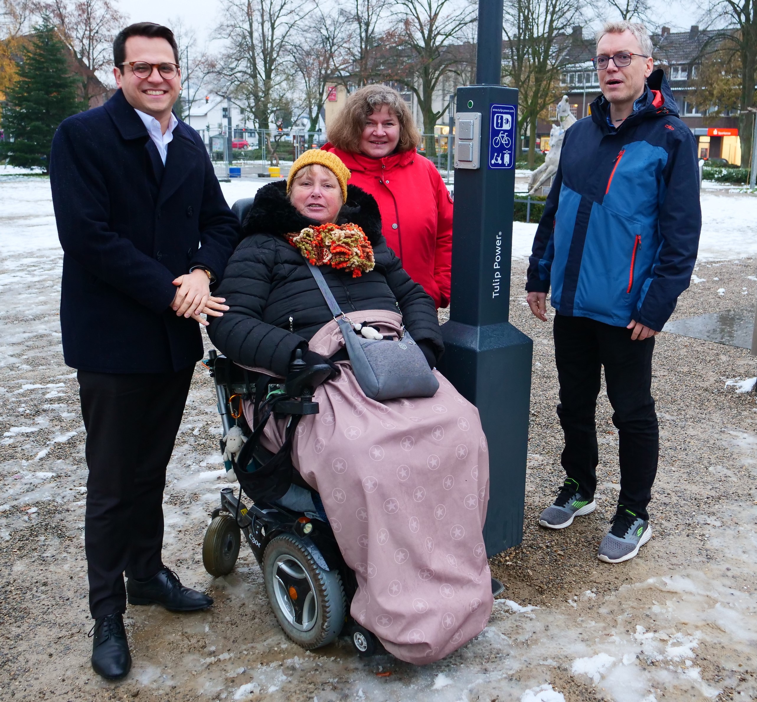 Vier Menschen (Herzogenraths Bürgermeister Dr. Fadavian, Gerrit Heidenreich vom Tiefbauamt, Elke Hofmann und Sabine Früke vom Forum für Menschen mit Behinderung Herzogenrath) in winterlicher Kleidung auf dem mit Schnee bedeckten August-Schmidt-Platz in Merkstein, neben der neuen Ladesäule stehend und lächelnd