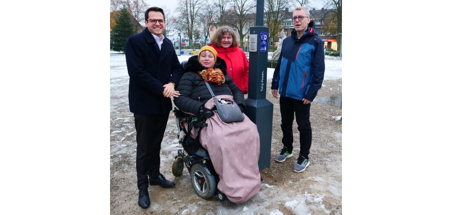 Vier Menschen (Herzogenraths Bürgermeister Dr. Fadavian, Gerrit Heidenreich vom Tiefbauamt, Elke Hofmann und Sabine Früke vom Forum für Menschen mit Behinderung Herzogenrath) in winterlicher Kleidung auf dem mit Schnee bedeckten August-Schmidt-Platz in Merkstein, neben der neuen Ladesäule stehend und lächelnd