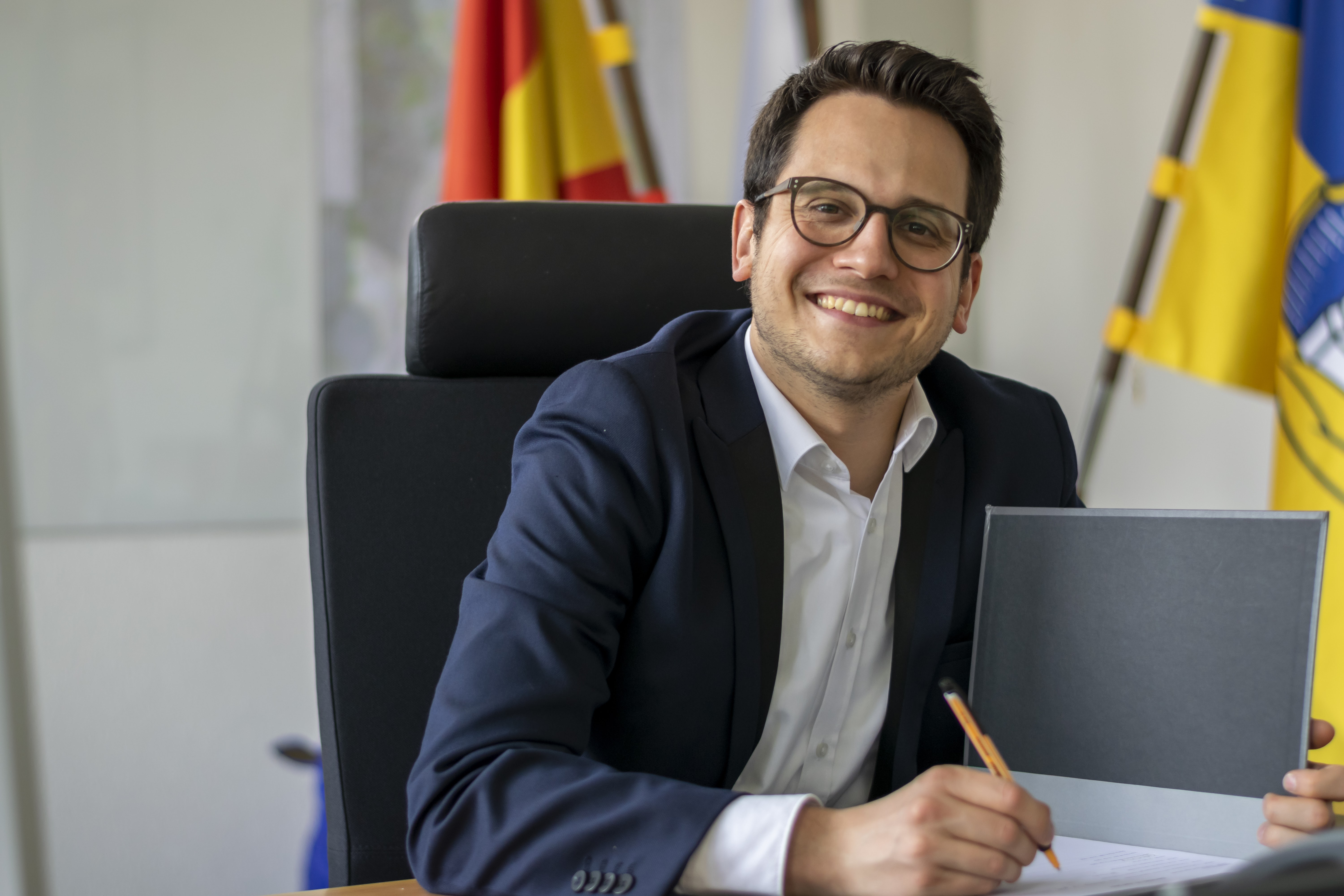 Herzogenraths Bürgermeister Dr. Benjamin Fadavian sitzt an seinem Schreibtisch und blickt lächelnd in die Kamera