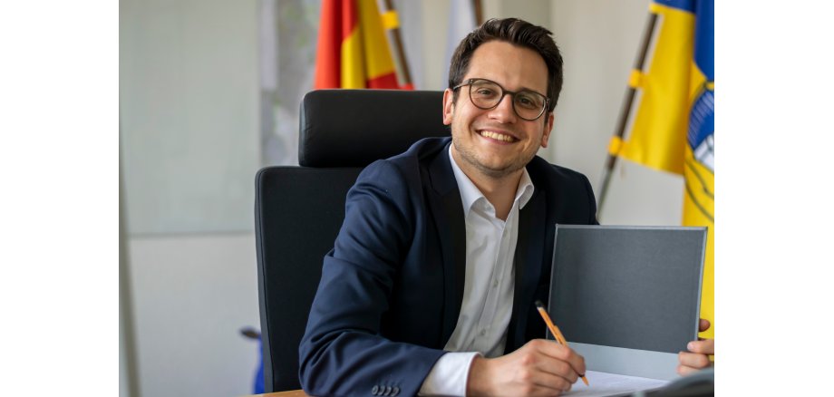 Herzogenraths Bürgermeister Dr. Benjamin Fadavian sitzt an seinem Schreibtisch und blickt lächelnd in die Kamera