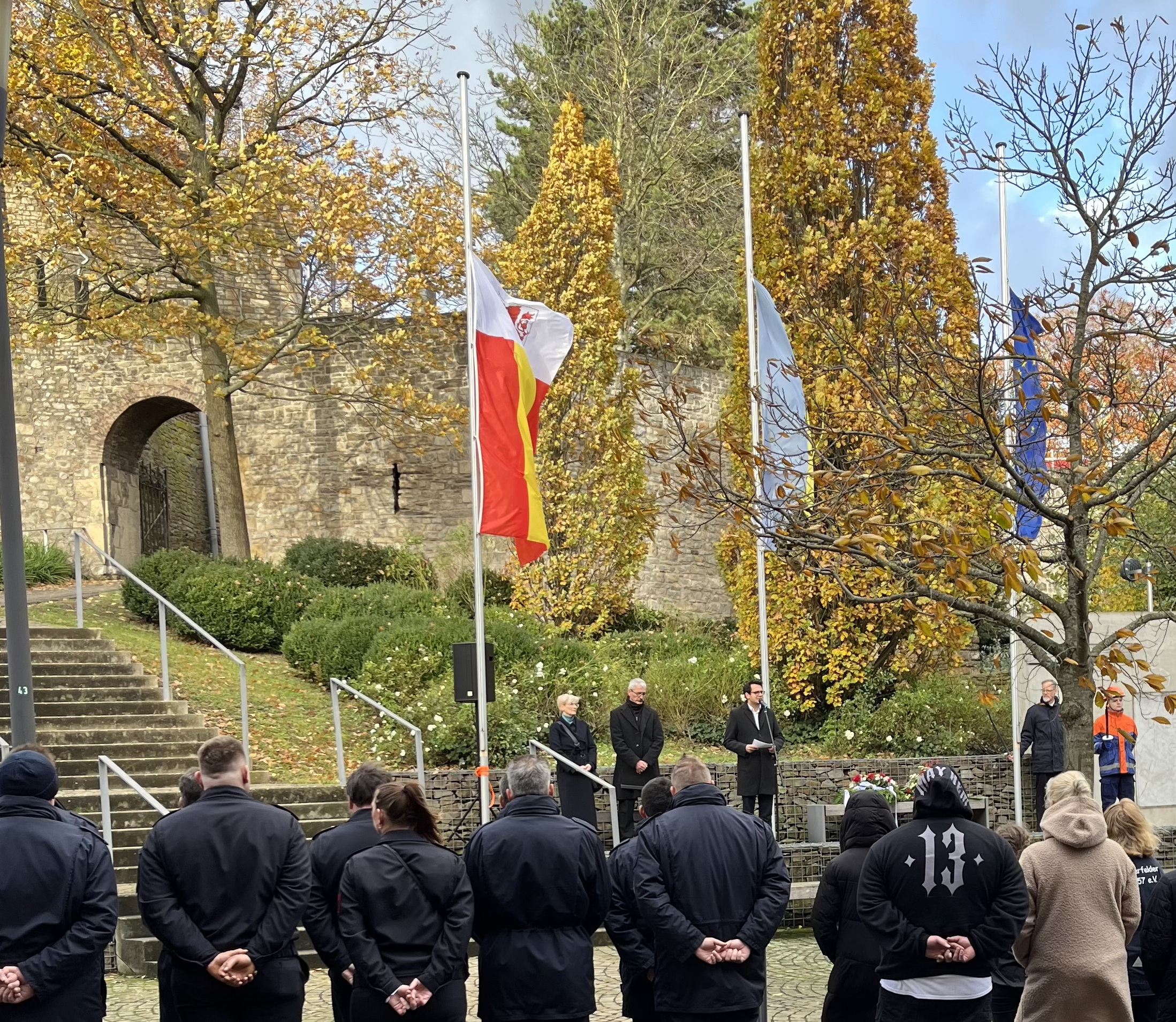 Herzogenraths Bürgermeister Dr. Benjamin Fadavian mit der Bürgermeisterin Kerkrades und dem Pastoralreferanten bei der Gedenkveranstaltung in Herzogenrath-Mitte an der Burg Rode, Flaggen auf Halbmast im Hintergrund