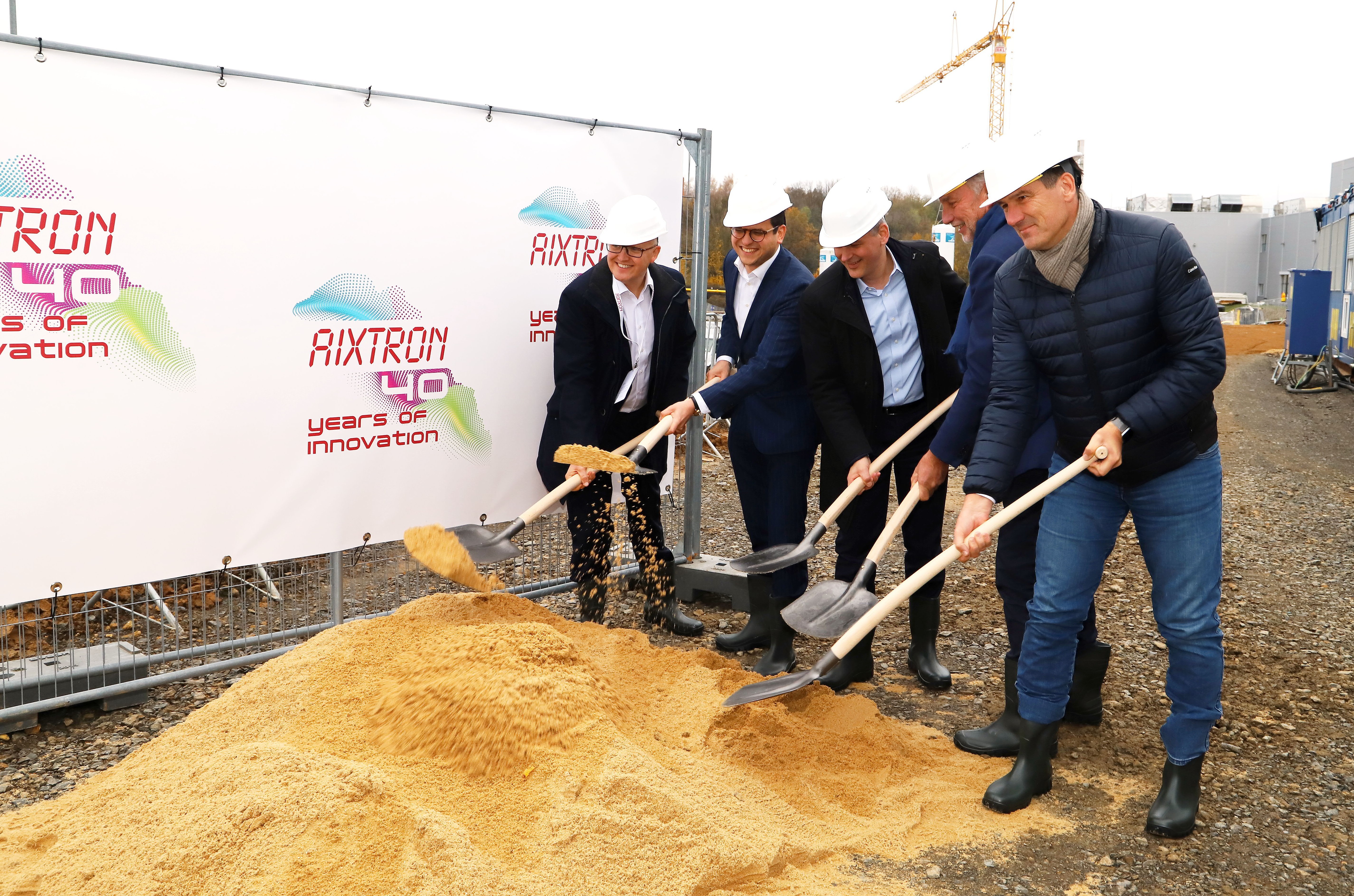 Fünf Männer (AIXTRON-Vorstand Dr. Christian Danninger, Herzogenraths Bürgermeister Dr. Benjamin Fadavian, AIXTRON-Vorstand Dr. Felix Grawert, AIXTRON-Aufsichtsratsvorsitzender Kim Schindelhauer und Herzogenraths Technischer Beigeordneter Franz-Josef Türck-Hövener) mit Helmen und Schaufeln voller Sand bei der Zeremonie zum Spatenstich des neuen Innovationszentrums der Firma AIXTRON in Herzogenrath