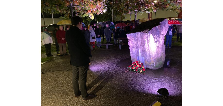 Abgebildet ist Herzogenraths Bürgermeister Dr. Benjamin Fadavian, wie er mit dem Rücken zur Kamera vor dem Mahnmal am Herzogenrather Rathaus steht, nachdem er einen Kranz für die Opfer des NS-Terrors niedergelegt hat. Um ihn und das Mahnmal herum stehen zahlreiche Besucher mit Regenschirmen