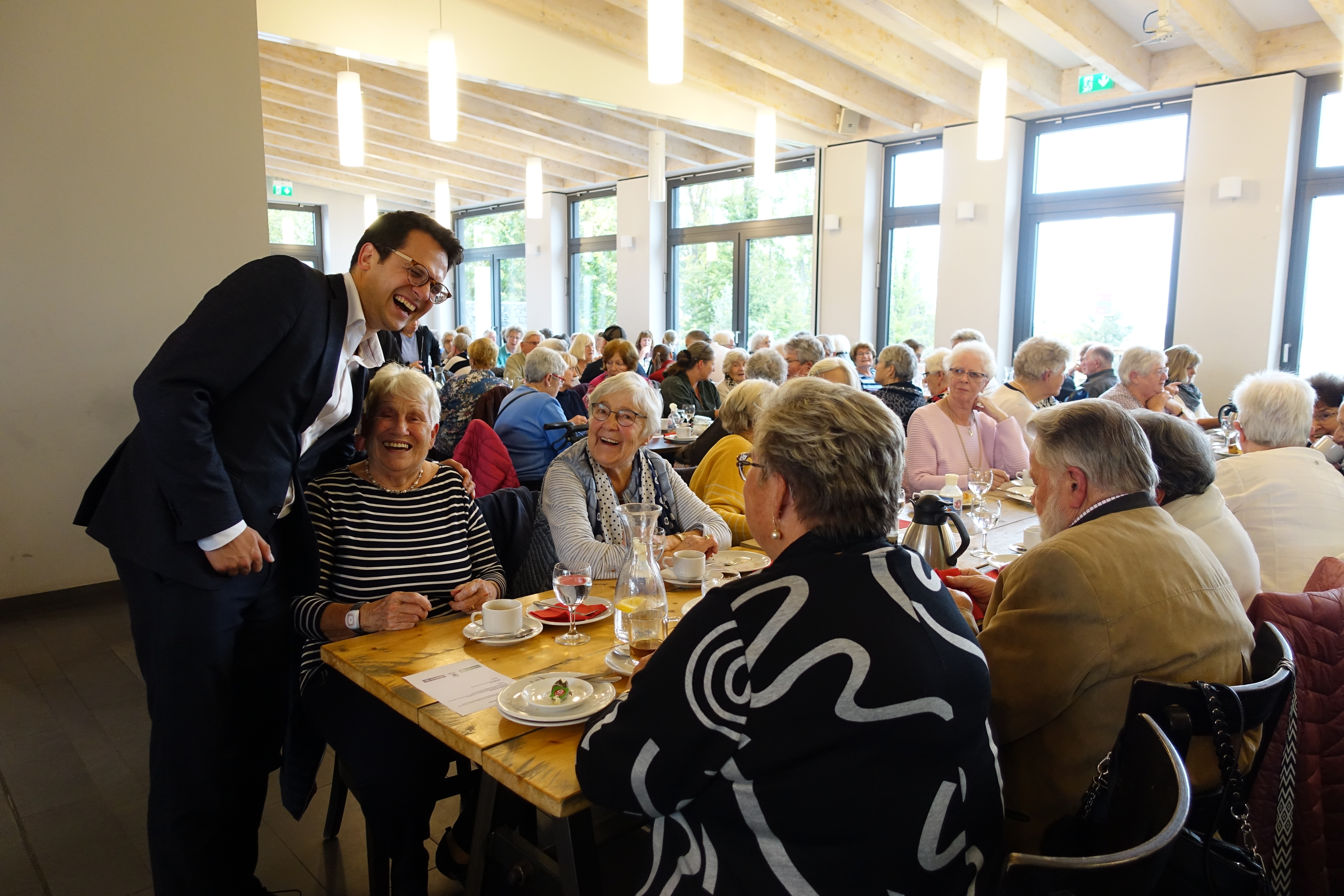 Abgebildet sind Herzogenraths Bürgermeister Dr. Benjamin Fadavian und Teilnehmer der Seniorenfahrt nach Inden: Der Bürgermeister besuchte die Senioren, als diese gerade im Restaurant aßen. Die Menschen auf dem Bild lachen