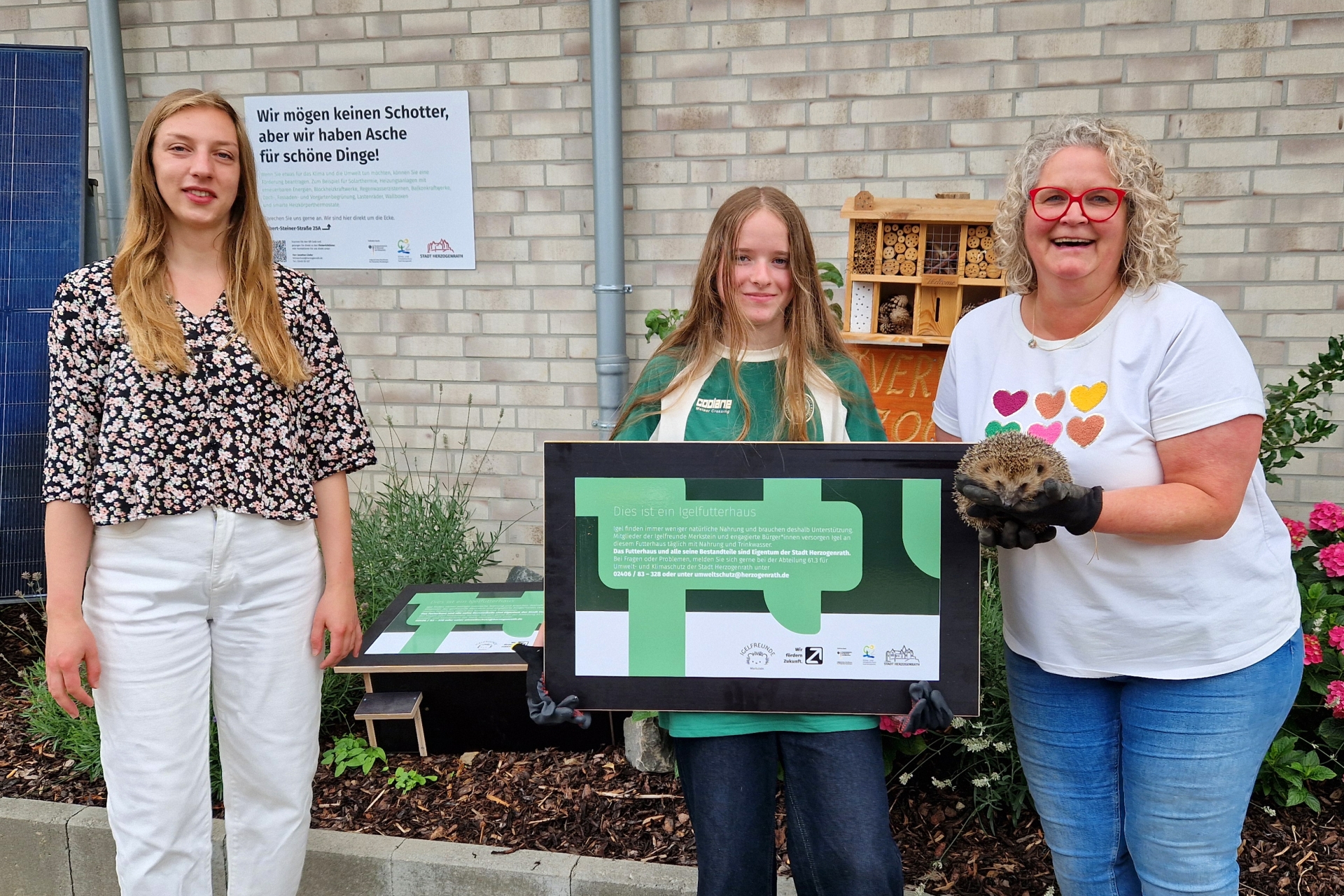 Projektleiterin Fabienne Heinrichs von der Umwelt- und Klimaschutzabteilung der Stadt Herzogenrath, Kinderbürgermeisterin Zoe Kißling und Sandra Nitschke von den Igelfreunden Merkstein mit dem aufgepäppelten Igel „Olaf“. Im Hintergrund ist eine zusammengebaute Igelfutterstation im Pflanzbeet der Umwelt- und Klimaschutzabteilung an der Wurmresidenz zu sehen.