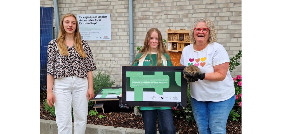 Projektleiterin Fabienne Heinrichs von der Umwelt- und Klimaschutzabteilung der Stadt Herzogenrath, Kinderbürgermeisterin Zoe Kißling und Sandra Nitschke von den Igelfreunden Merkstein mit dem aufgepäppelten Igel „Olaf“. Im Hintergrund ist eine zusammengebaute Igelfutterstation im Pflanzbeet der Umwelt- und Klimaschutzabteilung an der Wurmresidenz zu sehen.