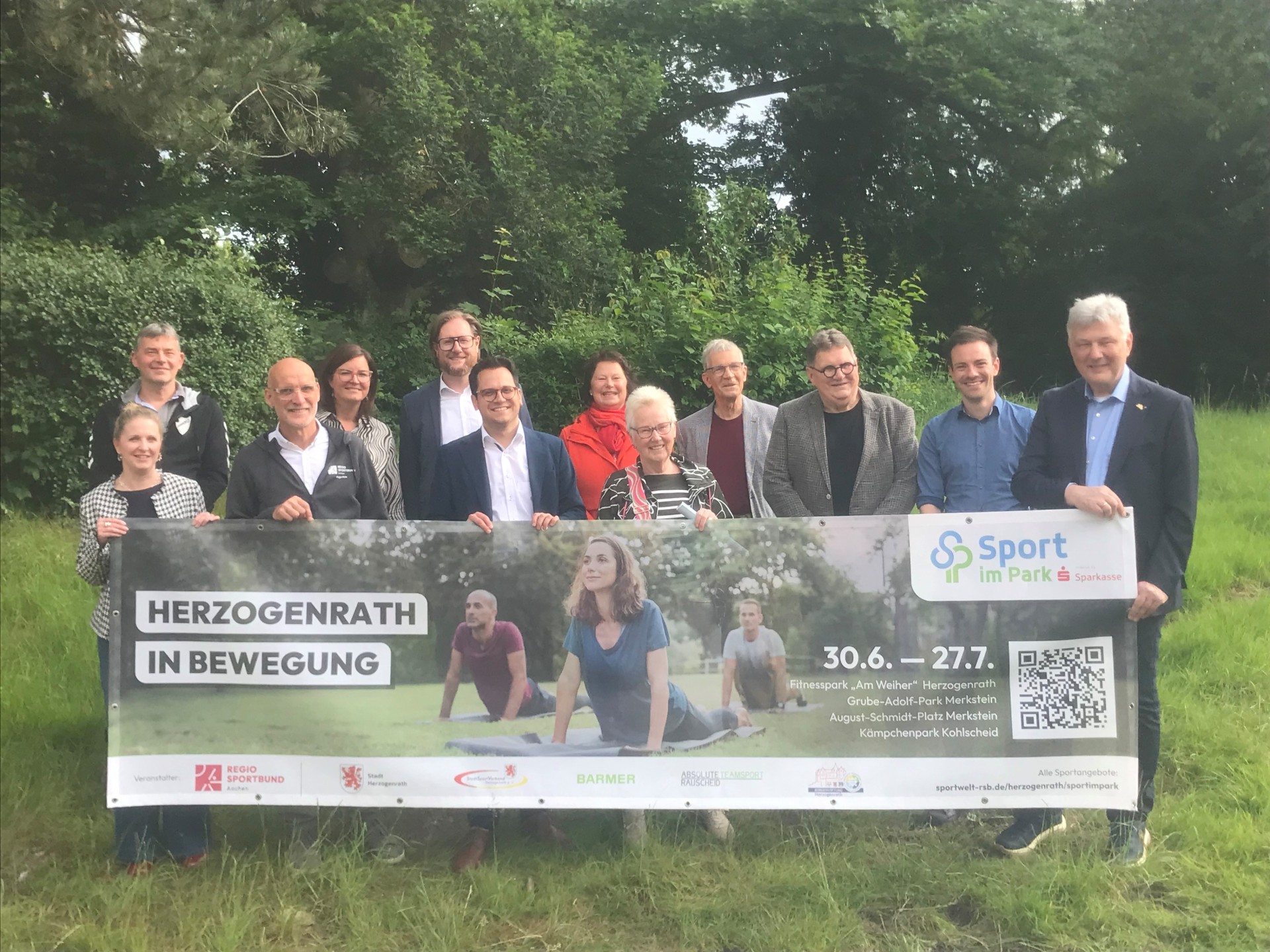 Die Sponsoren und Organisatoren von "Sport im Park" halten lächelnd zwischen Bäumen ein Banner mit Daten zur Aktion hoch - unter ihnen auch Bürgermeister Dr. Benjamin Fadavian