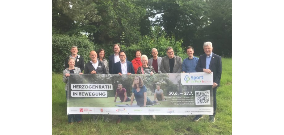 Die Sponsoren und Organisatoren von "Sport im Park" halten lächelnd zwischen Bäumen ein Banner mit Daten zur Aktion hoch - unter ihnen auch Bürgermeister Dr. Benjamin Fadavian