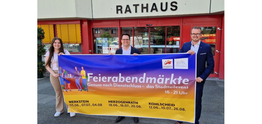 Das Bild zeigt v.l.n.r. Emily Nievelstein (Geschäftsführerin Stadtmarketing Herzogenrath e.V.), Bürgermeister Dr. Benjamin Fadavian und Yannick Wimmers (Büroleiter Bürgermeisterbüro) mit dem Ankündigungsbanner für die diesjährigen Feierabendmärkte